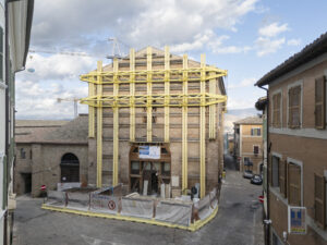 Edificio in mattoni con strutture gialle di supporto e cantiere recintato all'incrocio di strade urbane.