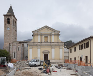 Cantiere davanti a chiesa gialla con torre campanaria in pietra e orologio, cielo nuvoloso e colline.