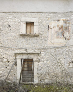 Facciata in pietra con porta sorretta da assi, finestra chiusa e affresco sbiadito a destra.