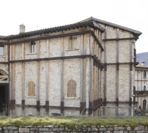 Edificio in pietra con finestre ad arco e rinforzi in legno e metallo su terreno con erba e muschio.