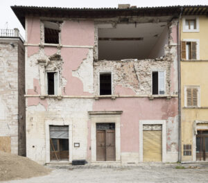 Facciata di edificio danneggiato con pareti e finestre crollate e macerie davanti.