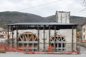 Struttura storica in pietra con archi in legno, recintata e coperta da tetto metallico in contesto urbano.