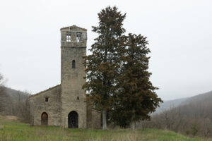 Edificio in pietra con torre campanaria accanto a due alberi in campo verde e colline sullo sfondo.