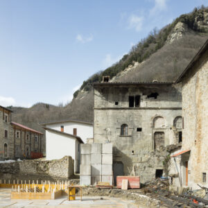 Cantiere con strutture in cemento davanti a edificio antico in pietra e colline alberate sullo sfondo.