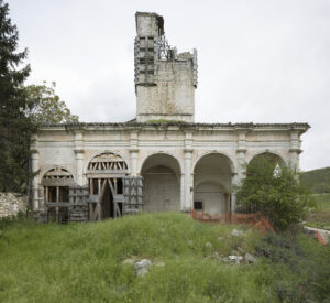 Resti di edificio storico in pietra con archi e rinforzi in legno e metallo, area verde e recinzione arancione.