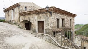 Casa in pietra con tetto in tegole e strada inclinata in contesto rurale con colline verdi.