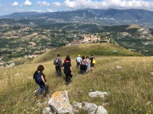 Persone che camminano su sentiero erboso verso paese su collina con montagne sullo sfondo.