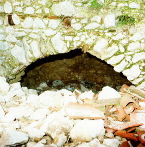 Apertura ad arco in muro di pietra con detriti e frammenti di mattoni davanti all'ingresso.
