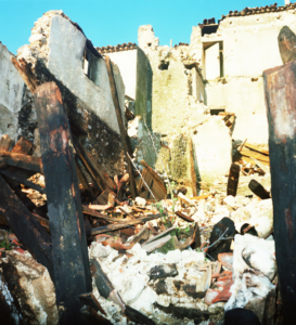 Rovine di edificio crollato con travi di legno bruciate e macerie sotto cielo sereno.