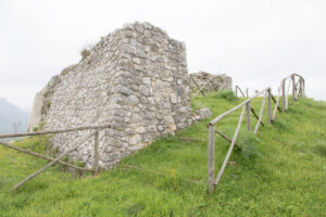 Ruderi di muro in pietra con recinzione in legno su area erbosa e collinare.