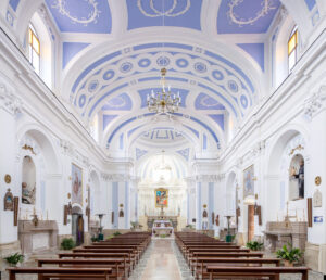 Interno di chiesa con soffitto blu decorato, grandi finestre, banchi e altare maggiore sul fondo.