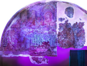Affresco semicircolare danneggiato con scena religiosa di figure sedute attorno a tavola imbandita.