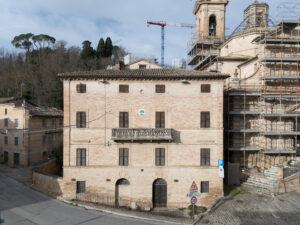 Edificio in mattoni con persiane chiuse e balcone centrale, accanto a chiesa in ristrutturazione con impalcature.