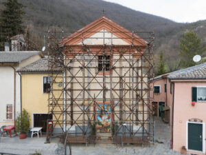 Facciata di una chiesa in ristrutturazione con impalcature tra case colorate e colline sullo sfondo.