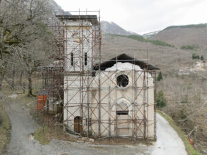 Chiesa in pietra con impalcatura per restauro in area montuosa con alberi spogli e colline.