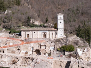 Chiesa in pietra con campanile su collina davanti a ruderi delimitati da recinzione arancione.