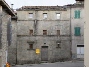 Edificio storico in pietra con finestre rettangolari, porta in legno e persiane verdi su strada asfaltata.