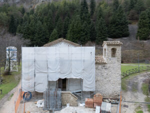 Piccola chiesa in pietra in restauro con impalcature e materiali edili, bosco sullo sfondo.