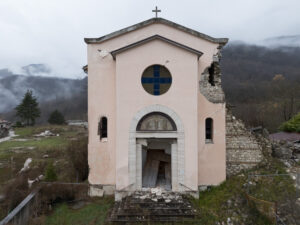 Chiesa danneggiata con facciata lesionata e muro crollato in area rurale con colline.
