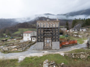 Piccola chiesa in pietra con impalcatura metallica in un'area rurale con nebbia e colline sullo sfondo.