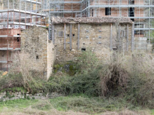 Vecchio edificio in pietra con tetto in tegole e impalcature, vegetazione e nuovo edificio in costruzione.