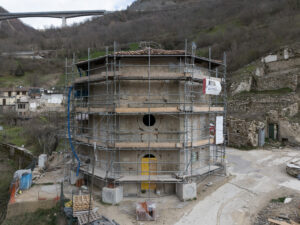 Edificio circolare in pietra con impalcature in un'area collinare con resti murari e viadotto sullo sfondo.
