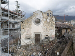 Facciata parzialmente crollata di edificio in pietra con detriti, impalcature e montagne sullo sfondo.