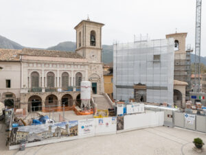 Cantiere di restauro davanti a due edifici storici con torri campanarie e montagne sullo sfondo