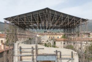 Struttura metallica con tetto sopra rovine di edificio antico, case e colline sullo sfondo.