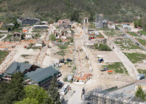 Cantiere di restauro con edifici parzialmente demoliti, ponteggi, mezzi e operai, in area collinare.