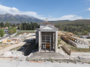 Edificio in pietra con impalcature in un cantiere, montagne innevate e boschi sullo sfondo.