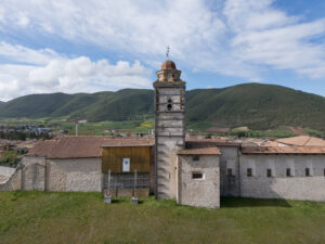 Antica struttura in pietra con campanile, tetti in tegole rosse, colline verdi e cielo nuvoloso.
