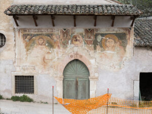 Facciata di edificio storico con portone ad arco e affreschi religiosi deteriorati con rete arancione davanti
