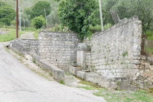 Fontana in pietra e muro lungo strada rurale con vegetazione e colline verdi.