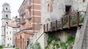 Edificio storico in pietra accanto a struttura moderna in mattoni con balconi e torre campanaria sullo sfondo.