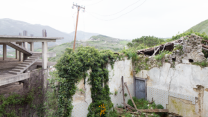Ruderi di vecchia costruzione con vegetazione e edificio incompleto in cemento su paesaggio collinare.