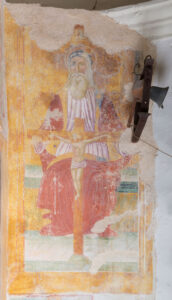 Affresco religioso consunto con Dio Padre anziano e crocifisso, campana metallica sul muro.