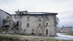 Casa in pietra parzialmente crollata con danni evidenti, strada asfaltata e edificio con torre campanaria sullo sfondo.