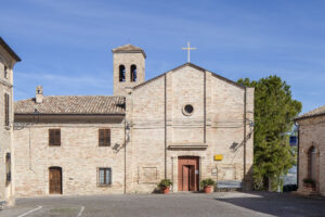 Chiesa in mattoni chiari con portone e rosone, campanile, panchina e alberi sotto cielo sereno.