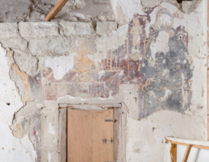 Parete con affresco parzialmente deteriorato e porta in legno con tracce di pittura murale.