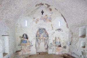 Interno di cappella con affreschi antichi parzialmente deteriorati e due strette finestrelle luminose.