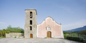 Chiesa con facciata rosa e torre campanaria grigia in area con pavimentazione di pietra.