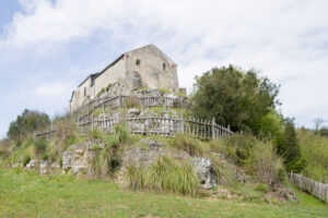 Costruzione antica in pietra su collina rocciosa con recinzione di legno e vegetazione intorno.
