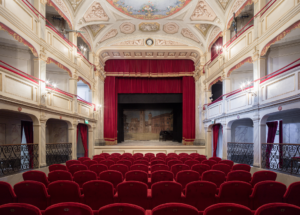 Interno teatro storico con poltrone rosse, balconate decorate, soffitto affrescato e palco con sipario rosso.