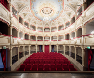 Interno di teatro storico con file di poltrone rosse, balconate decorate e soffitto affrescato.