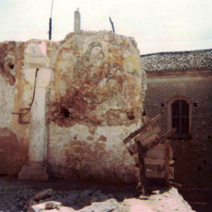 Parete in rovina con affreschi sacri, colonna isolata e edificio con finestra ad arco.