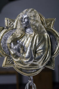 Medaglione metallico con figura scolpita di persona con capelli lunghi e mani intrecciate sul petto.