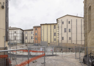 Area recintata con recinzioni metalliche e nastro arancione davanti a edifici storici.