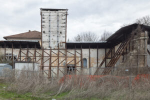 Edificio degradato con supporti metallici esterni, recinzioni e vegetazione secca circostante.