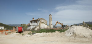 Cantiere con escavatore, rovine di edifici e campanile in paesaggio collinare soleggiato.